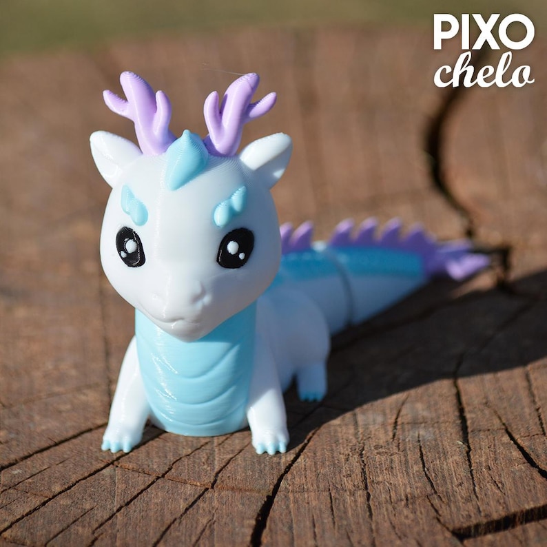 Peut inclure: Figurine de dragon blanc et bleu clair avec des d&eacute;tails violets. Le dragon a de grands yeux noirs, de petites cornes violettes et un corps segment&eacute;. La figurine est pos&eacute;e sur une surface en bois, avec le texte "PIXO chelo" en haut &agrave; droite.