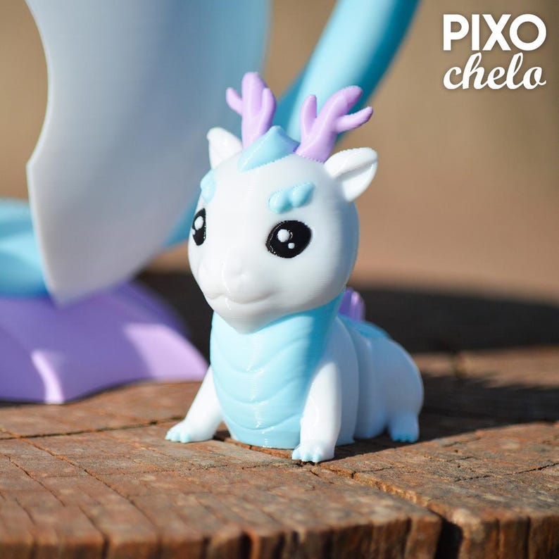 Peut inclure: Figurine de dragon blanc et bleu clair avec des bois violets. Le dragon a de grands yeux noirs et est assis sur une surface en bois. La figurine a une poitrine et un dos bleu clair. Le texte "PIXO chelo" est dans le coin sup&eacute;rieur droit.