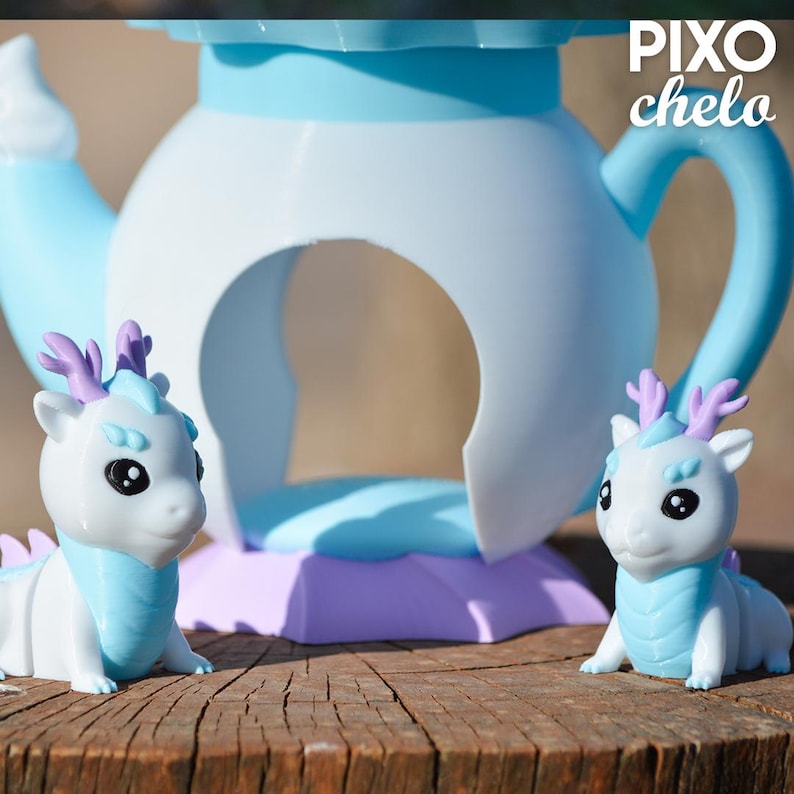 Peut inclure: Deux figurines de dragon blanc et bleu avec des bois violets sont pos&eacute;es sur une surface en bois devant un objet en forme de th&eacute;i&egrave;re bleu clair et blanc. Les figurines de dragon ont de grands yeux noirs et des accents bleus. La th&eacute;i&egrave;re a une grande ouverture et une anse.