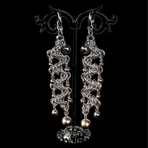 May include: Silver-toned chainmail earrings with a dangling design. The earrings feature a series of interconnected links and small beads, culminating in larger spherical accents. The earrings are displayed on a black stand against a black background.