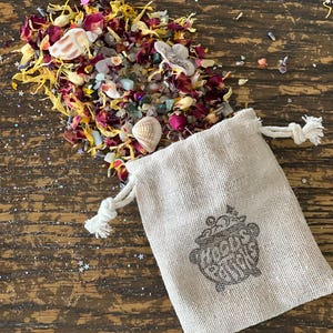May include: A beige drawstring bag with a cauldron graphic and the text "Magic Potions" is next to a pile of dried flowers, seashells, and small crystals. The flowers are yellow, red, and white.