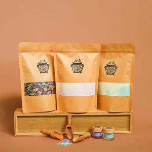 Pode incluir: Três sacos de papel castanho com o texto "Hocus Potion" e um logótipo de caldeirão. Os sacos são preenchidos com ingredientes coloridos. Há colheres de madeira e pequenos frascos de pó colorido à frente. O fundo é de cor pêssego lisa.
