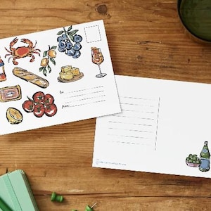 May include: Two white postcards featuring colorful hand-drawn illustrations of food and drink items including a crab, baguette, tomatoes, cheese, blueberries, and a cocktail. One postcard has lines for mailing addresses, while the other shows a small bowl of olives.
