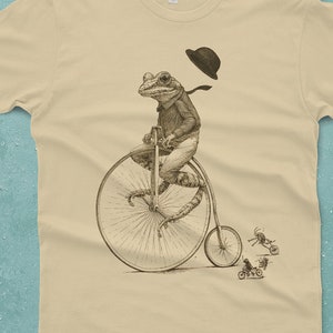 May include: A beige t-shirt with a graphic of a frog wearing a hat and riding a penny-farthing bicycle. The frog is wearing a bow tie and has a long tail. There are smaller insects riding bicycles in the background.
