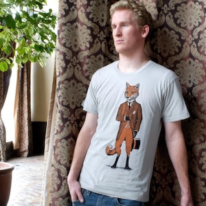May include: A light gray t-shirt featuring a dapper fox illustration. The fox is depicted in a brown suit, orange pants, and a top hat, holding a briefcase. The shirt is paired with blue jeans.
