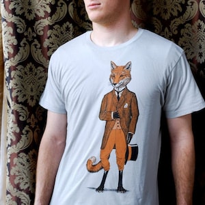 Puede incluir: Una camiseta gris claro con un gráfico de un zorro con traje y sombrero de copa. El zorro es naranja y blanco con una cola negra.