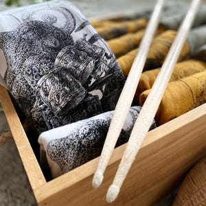 A box full of T-shirts with octopuses playing drums on them. They are light gray, gold and light green. There are drum sticks on top of all of them.