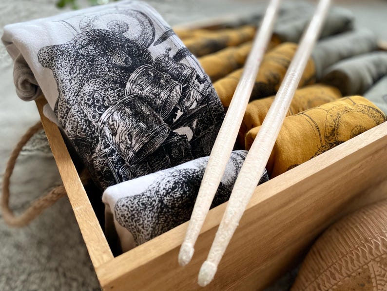 A box full of T-shirts with octopuses playing drums on them. They are light gray, gold and light green. There are drum sticks on top of all of them.