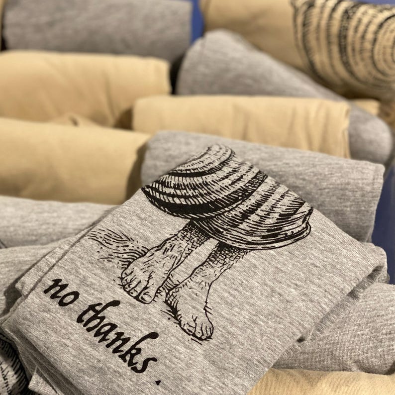 A pile of folded shirts with the image of a shell that has human feet sticking out of it. There is a caption under it that reads "no thanks". It's printed in black onto heather gray and cream T-shirts.