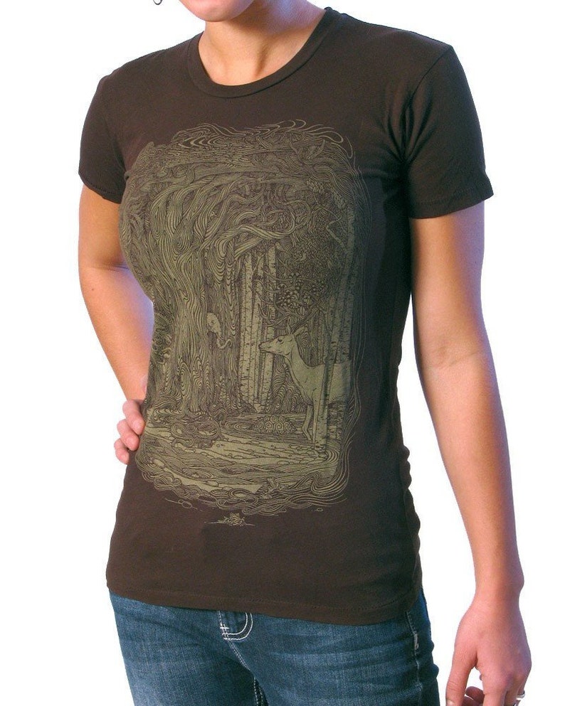 Puede incluir: Una camiseta marr&oacute;n con un estampado gr&aacute;fico verde y marr&oacute;n de una escena de bosque con un ciervo. El estampado gr&aacute;fico es detallado e intrincado.