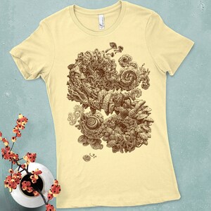 Op de afbeelding: Lichtgele t-shirt met een sepia-getinte illustratie van een abstract, organisch ontwerp. Het kunstwerk bevat wervelende vormen, paddenstoelachtige vormen en ingewikkelde texturen. Het shirt heeft een klassieke ronde hals en korte mouwen.