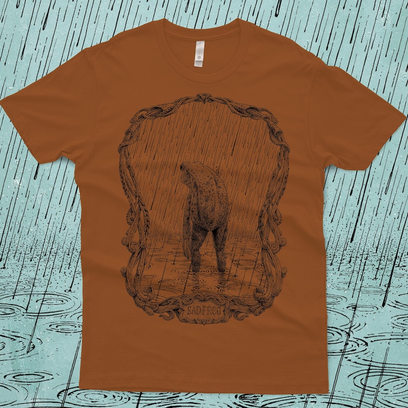A drawing of a sad frog standing in the rain, surrounded by an ornate border. Screen printed onto a clay, Next Level brand T-shirt.