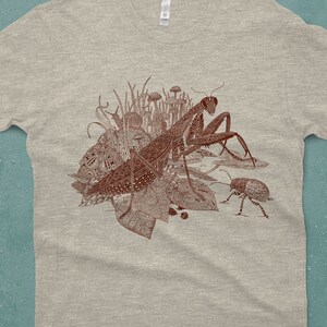 An intricate drawing of a praying mantis and other insects and creatures in front of mushrooms, leaves and grass. It is screen printed on a heather gray Next Level brand T-shirt in front of a turquoise backdrop