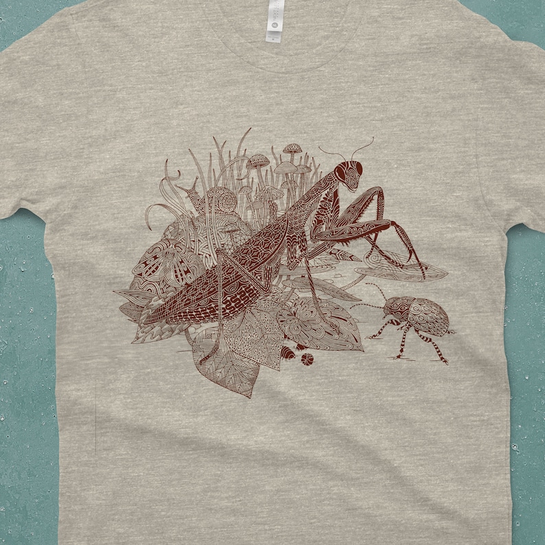 An intricate drawing of a praying mantis and other insects and creatures in front of mushrooms, leaves and grass. It is screen printed on a heather gray Next Level brand T-shirt in front of a turquoise backdrop