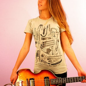 Puede incluir: Una mujer con una camiseta gris claro con una ilustraci&oacute;n en blanco y negro de instrumentos musicales. Est&aacute; sosteniendo un bajo el&eacute;ctrico marr&oacute;n y naranja.