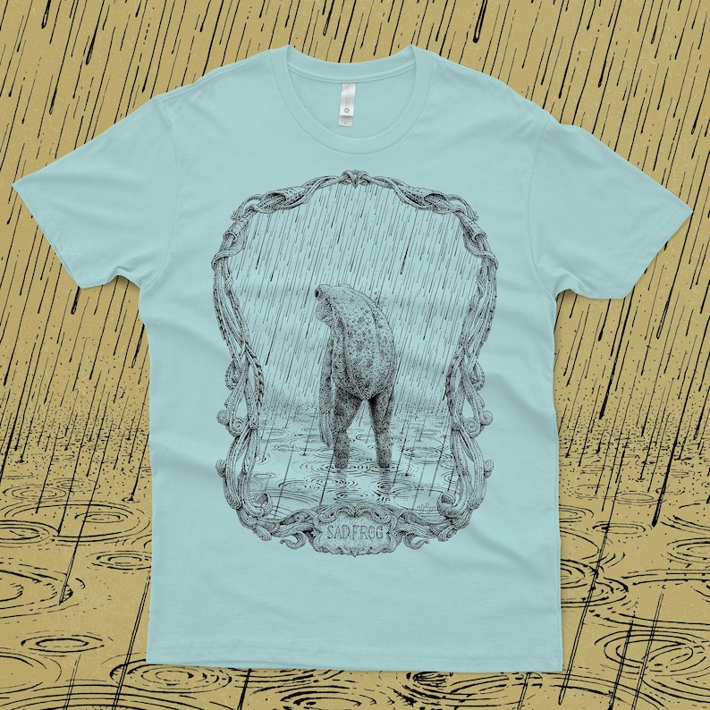 A drawing of a sad frog standing in the rain, surrounded by an ornate border. Screen printed onto a blue, Next Level brand T-shirt.