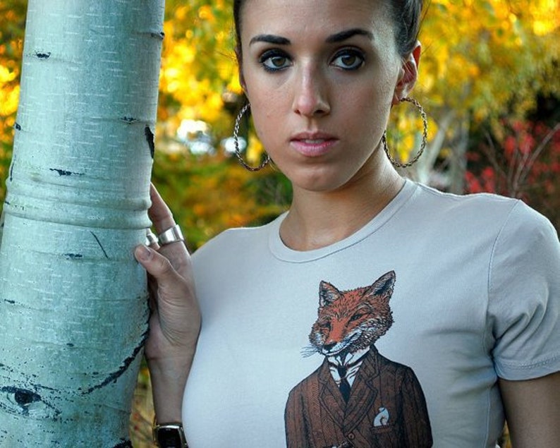 Puede incluir: Una mujer lleva una camiseta gris claro con un gr&aacute;fico de un zorro con traje y corbata. El zorro es marr&oacute;n y naranja con un cuello blanco y una pajarita negra. La mujer est&aacute; de pie junto a un &aacute;rbol con corteza blanca.