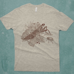 An intricate drawing of a praying mantis and other insects and creatures in front of mushrooms, leaves and grass. It is screen printed on a heather gray unisex or men's T-shirt in front of a turquoise backdrop