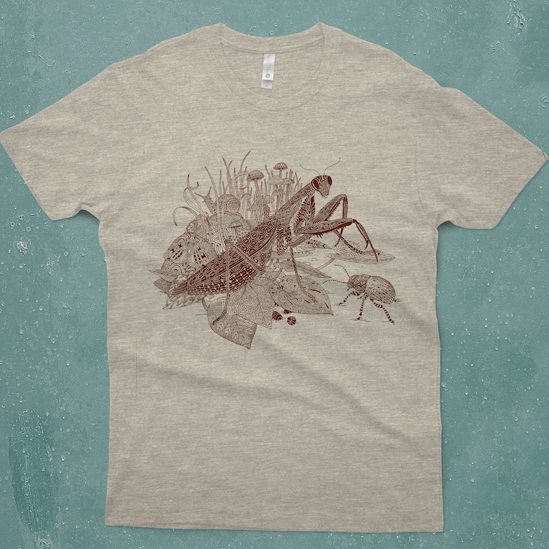 An intricate drawing of a praying mantis and other insects and creatures in front of mushrooms, leaves and grass. It is screen printed on a heather gray unisex or men's T-shirt in front of a turquoise backdrop
