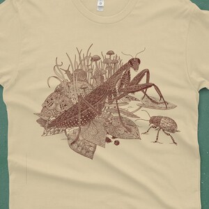 An intricate drawing of a praying mantis and other insects and creatures in front of mushrooms, leaves and grass. It is screen printed on a cream colored unisex-mens T-shirt in front of a turquoise backdrop
