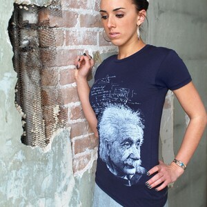 Puede incluir: Una mujer lleva una camiseta azul marino con un gr&aacute;fico blanco de Albert Einstein. La camiseta tiene una impresi&oacute;n blanca de ecuaciones y f&oacute;rmulas matem&aacute;ticas. La mujer est&aacute; de pie frente a una pared de ladrillo.