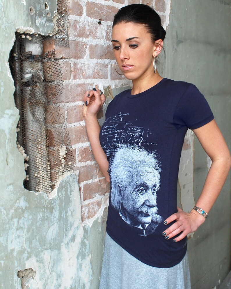 Puede incluir: Una mujer lleva una camiseta azul marino con un gr&aacute;fico blanco de Albert Einstein. La camiseta tiene una impresi&oacute;n blanca de ecuaciones y f&oacute;rmulas matem&aacute;ticas. La mujer est&aacute; de pie frente a una pared de ladrillo.