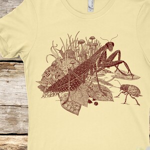 May include: A maroon-colored illustration of a praying mantis, mushrooms, and other insects on a light yellow t-shirt.