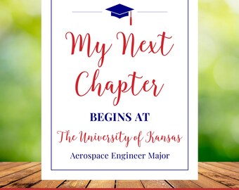 May include: A white sign with a blue border and text in red and blue. The text reads "My Next Chapter Begins At The University of Kansas Aerospace Engineer Major". A graduation cap graphic is at the top.
