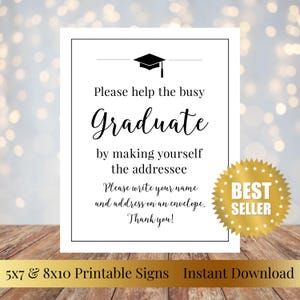 May include: A white sign with a black border and text that reads "Please help the busy Graduate." Includes a graduation cap graphic and a "Best Seller" sticker. The sign is on a wooden surface.