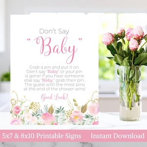 Può includere: Un cartello bianco con la scritta "Don't Say Baby" in rosa. Il cartello include le istruzioni per un gioco per il baby shower, con accenti floreali e dorati. Un vaso di fiori rosa è a destra.