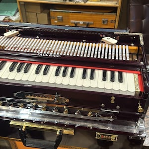 May include: A dark burgundy harmonium with a keyboard, reeds, and the text "PAUL & CO." The instrument has a red accent and gold-colored hardware. It is a traditional musical instrument.