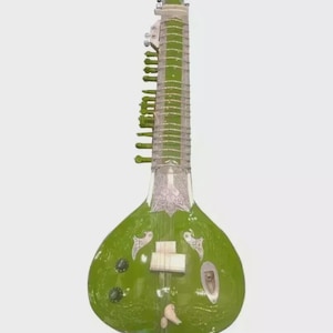 May include: A green sitar, a long-necked stringed instrument, stands against a white background. The instrument features a pear-shaped body, a long neck with frets, and multiple strings. Decorative silver-colored accents adorn the body.