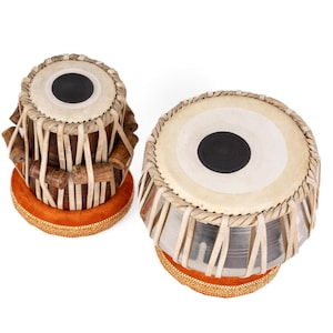 May include: A pair of tabla drums featuring a smaller wooden dayan and a larger metallic bayan. Both drums have white skin heads with central black circular patches, secured by light-colored leather tension straps and resting on orange rings.