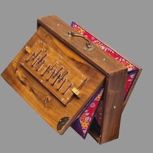 May include: A wooden harmonium with a brass handle and decorative fabric lining. The instrument has a keyboard-like section with metal pins and a wooden frame. The fabric features a floral pattern with red, purple, and yellow hues.