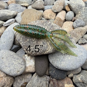 May include: A green and clear fishing lure with orange and green speckles, measuring 4.5 inches. The lure is designed to mimic a baitfish and has a segmented body and a forked tail. It is resting on a bed of rocks.