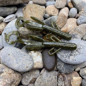 Peut inclure: Deux leurres de pêche en plastique souple vert et brun avec de multiples appendices. Les leurres sont posés sur un lit de petites pierres grises lisses.
