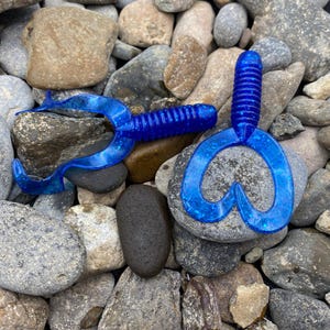 Peut inclure: Deux leurres de pêche en plastique bleu en forme de cœur avec un corps nervuré. Les leurres sont sur un lit de roches grises.