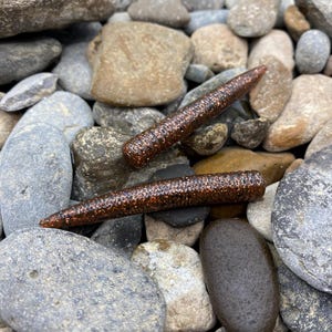 May include: Two cylindrical fishing lures with a dark brown, semi-translucent body embedded with fine copper-colored glitter. The lures feature a ribbed texture along their length and taper to a point at one end, resting on a bed of smooth stones.