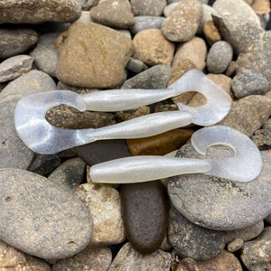 May include: Two translucent white plastic fishing lures with a worm shape and a curved tail. The lures are on a bed of gray and brown rocks.