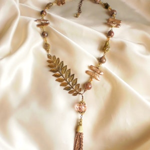 May include: A gold necklace with a leaf pendant, brown and clear beads, and a tassel. The necklace is on a white satin background.