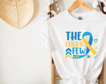 Può includere: Maglietta bianca con un nastro blu e giallo, il testo "THE LUCKY FEW 3.21" e altri piccoli elementi grafici.