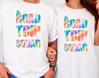 Op de afbeelding: Twee witte T-shirts met de tekst "ROAD TRIP SQUAD" in een kleurrijk tie-dye ontwerp. Beide personen dragen een blauwe jeans. Eén persoon heeft een mouwtattoo, de ander heeft lang haar.