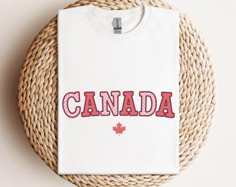 以下が含まれることがあります： 白いTシャツに、赤と白の文字で「CANADA」と書いてあり、その下に赤いカエデの葉が描かれています。