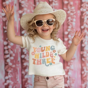 May include: A young girl wearing a white t-shirt with the text "Young Wild & Three" in colorful retro font and flowers. She is wearing a straw cowboy hat and sunglasses.