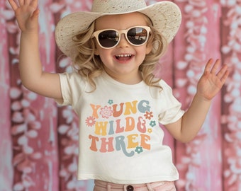 May include: A young girl wearing a white t-shirt with the text "Young Wild & Three" in colorful retro font and flowers. She is wearing a straw cowboy hat and sunglasses.