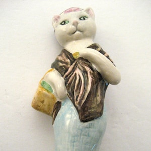 May include: A ceramic figurine of a cat-like figure wearing a brown jacket, blue pants, and pink shoes. The cat holds a small basket and has green eyes. The figurine is standing on a textured base.