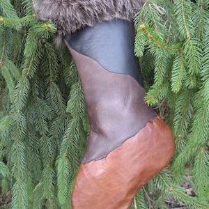 May include: A handmade Christmas stocking with a brown leather toe and body, a dark brown leather midsection, and a black leather upper. The top is trimmed with brown fur. The stocking hangs against a backdrop of green pine needles.