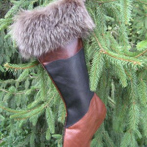May include: A Christmas stocking made of brown leather, black leather, and faux fur. The stocking is hanging from a tree branch. The top of the stocking is trimmed with brown faux fur.