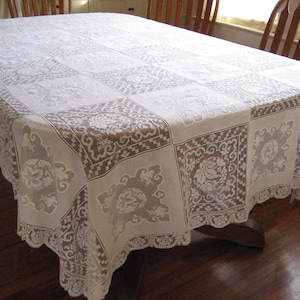 May include: A white lace tablecloth with a floral and geometric pattern covers a wooden table. The tablecloth has a scalloped edge and is draped over the table, showcasing its intricate design. The room has wooden chairs and a hardwood floor.