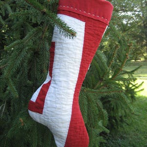 May include: A quilted Christmas stocking in red and white hangs from a pine branch. The stocking features a red cuff and heel, with a white central panel. The stitching is visible, and the stocking is filled with a soft material.
